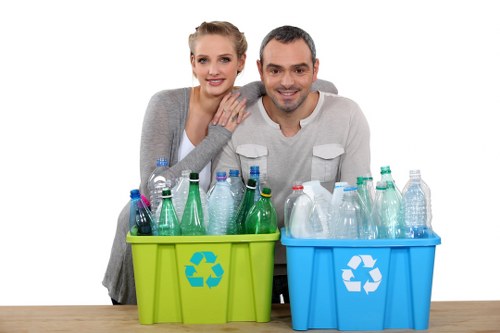 A smiling young couple, a woman with blonde hair and a man with short dark hair, sitting close together behind two plastic recycling bins on a wooden surface, one lime green and the other bright blue, each with a recycling symbol printed on the front. The green bin contains various used plastic bottles, some transparent and some with coloured labels, while the blue bin is filled with similar recyclable bottles. The background is plain white, and the lighting is bright and even, highlighting the clean, organized appearance of the recycling presentation. This image visually represents responsible waste segregation, aligning with services offered by Commercial Waste Removal Peckham in the South London area, focusing on rubbish and recyclable waste collection and disposal.