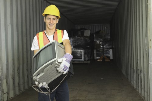 A young man wearing a yellow safety helmet, a high-visibility vest with orange and yellow reflective strips, and white gloves is standing inside a storage container or warehouse-like space. He is smiling and holding an older model computer monitor with a grey casing, which appears to be part of electronic waste. The background shows a dark interior with some black plastic-wrapped items or rubbish stacked towards the back of the space. The environment suggests a waste collection or disposal setting, typical of commercial rubbish removal services in Peckham, with a concrete or metal floor and corrugated metal walls. Natural light illuminates the scene, highlighting the man's attire, the computer monitor, and the surrounding industrial area, emphasizing the type of electronic waste handled by Commercial Waste Removal Peckham as part of their rubbish clearance operations in the area.