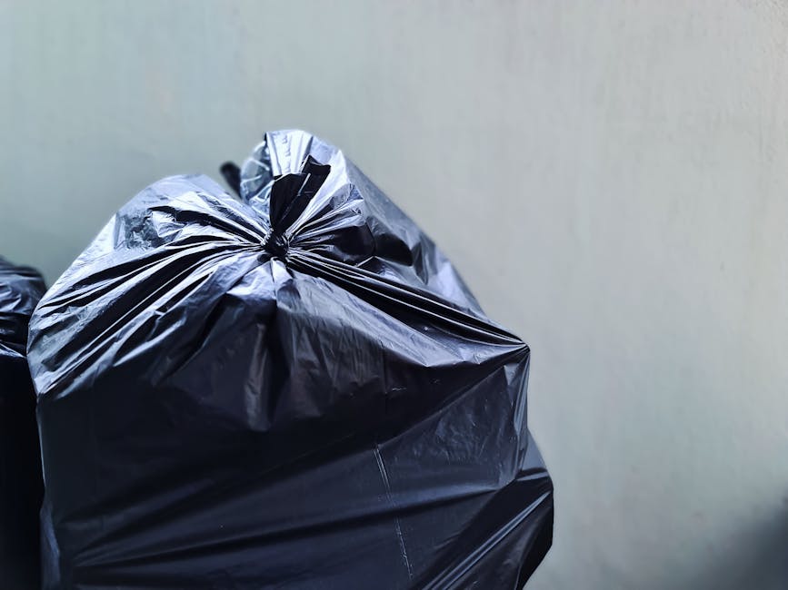 Close-up view of a large black plastic rubbish bag tied at the top, positioned in the foreground against a plain off-white wall background. The bag appears to be lightweight, slightly crinkled, and re